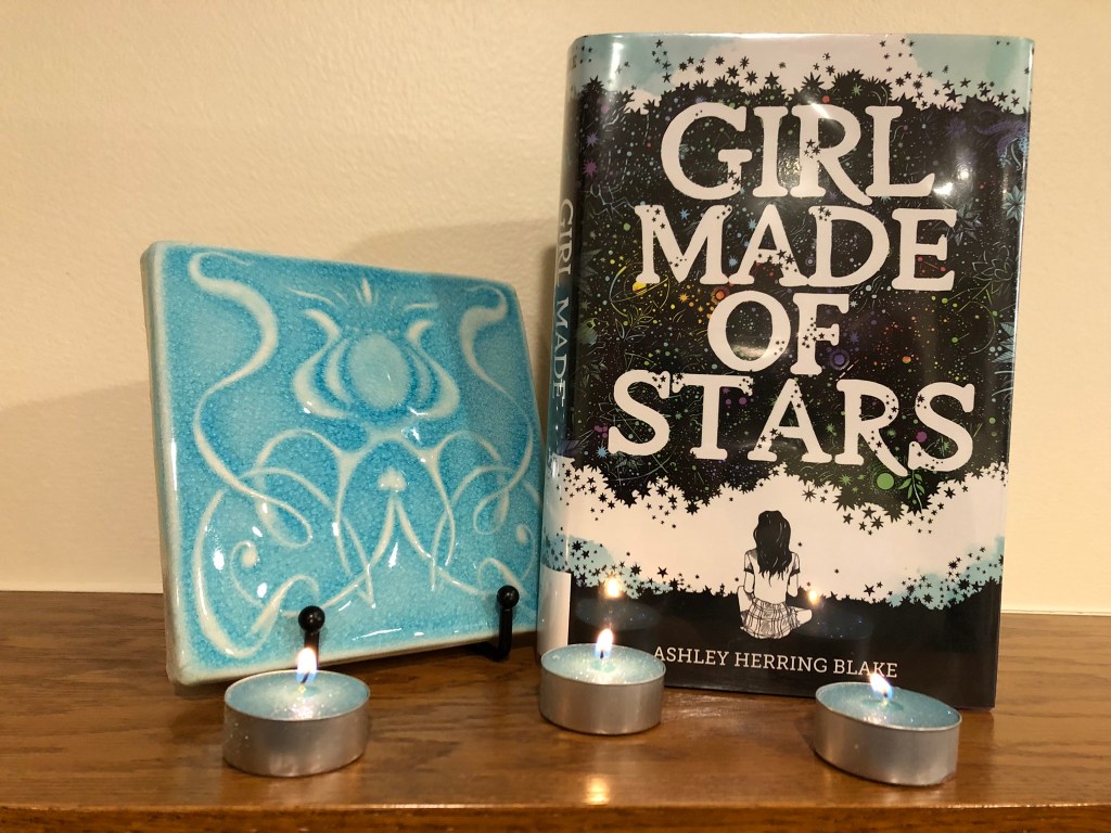 Image of book cover of Girl Made of Stars alongside a ceramic tile with an abstract design and three votive candles. 