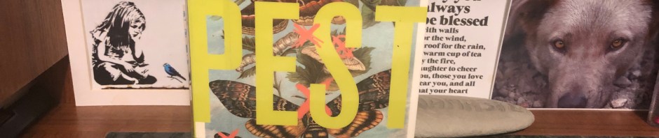 The book “Pest” standing on a desk with a few images behind it including a photo of the face of a wolfish dog. The book cover has images of bugs— a few butterflies, a few caterpillars, a few chrysalises. All have a red “X” over them.
